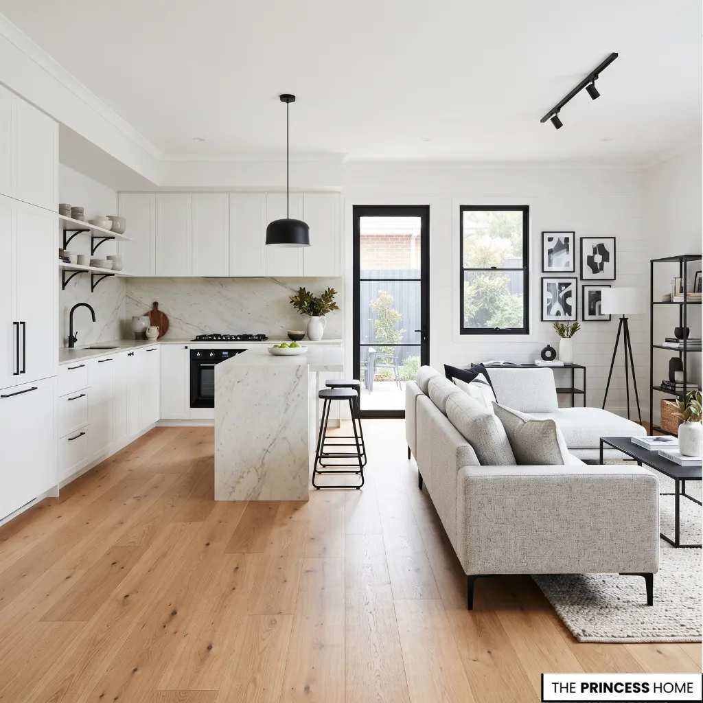 A bright, open-plan space featuring a modern white kitchen with a marble island and a cozy living area with a light gray sectional sofa and wooden flooring.