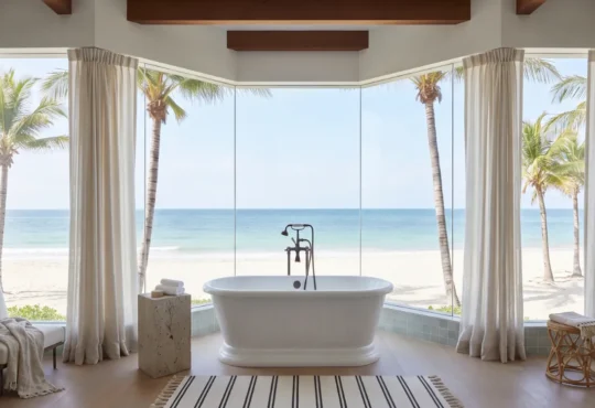 Luxury oceanfront bathroom featuring a freestanding tub, large panoramic windows, beach view, and palm trees.